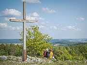 Zwei Wanderer sitzen neben dem Gipfelkreuz auf dem Bockberg und genießen die Aussicht