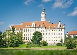 Großes, mehrstöckiges Gebäude mit orangefarbenem Dach und Turm, umgeben von Bäumen und Wiese, unter blauem Himmel.