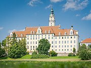 Großes, mehrstöckiges Gebäude mit orangefarbenem Dach und Turm, umgeben von Bäumen und Wiese, unter blauem Himmel.