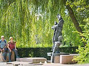 Zwei Personen sitzen auf einer Mauer neben einer abstrakten Skulptur im Freien, umgeben von Bäumen.