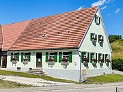 Weißes Haus mit grün lackierten Fensterläden und roten Blumen in Blumenkästen unter einem roten Ziegeldach bei blauem Himmel.